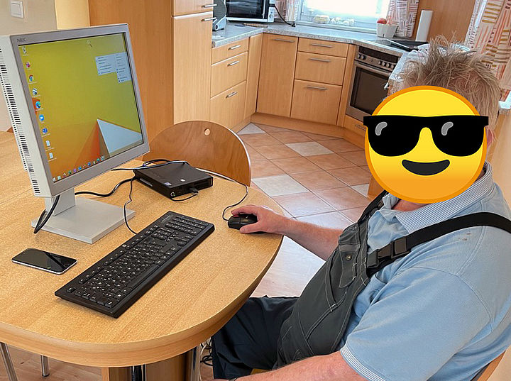 Ein Herr mit "Smiley-Gesicht" aus der Foto-Nachbearbeitung sitzt an einem Tisch mit der gespendeten PC-Ausrüstung und bedient die Computermaus.
