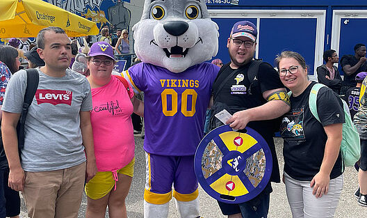 Zentral im Bild steht das Wolf-Maskottchen des American Footballteams „Vienna Vikings“ im lila-gelben Trikot. Links und rechts daneben posieren vier Teilnehmende und eine Betreuerin vor dem Stadion und lächeln in die Kamera.
