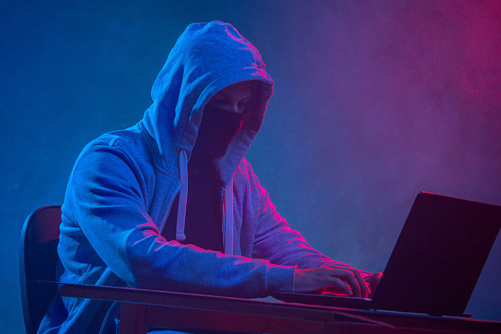 Ein vermummter Mann mit Kapuzenjacke sitzt im Dunkeln vor einem Notebook, die Hände auf der Tastatur.
