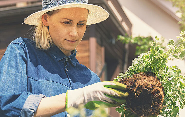 Eine blonde Frau mit Sonnenhut und Gartenhandschuhen. Sie steht im Garten und topft gerade eine Pflanze um.