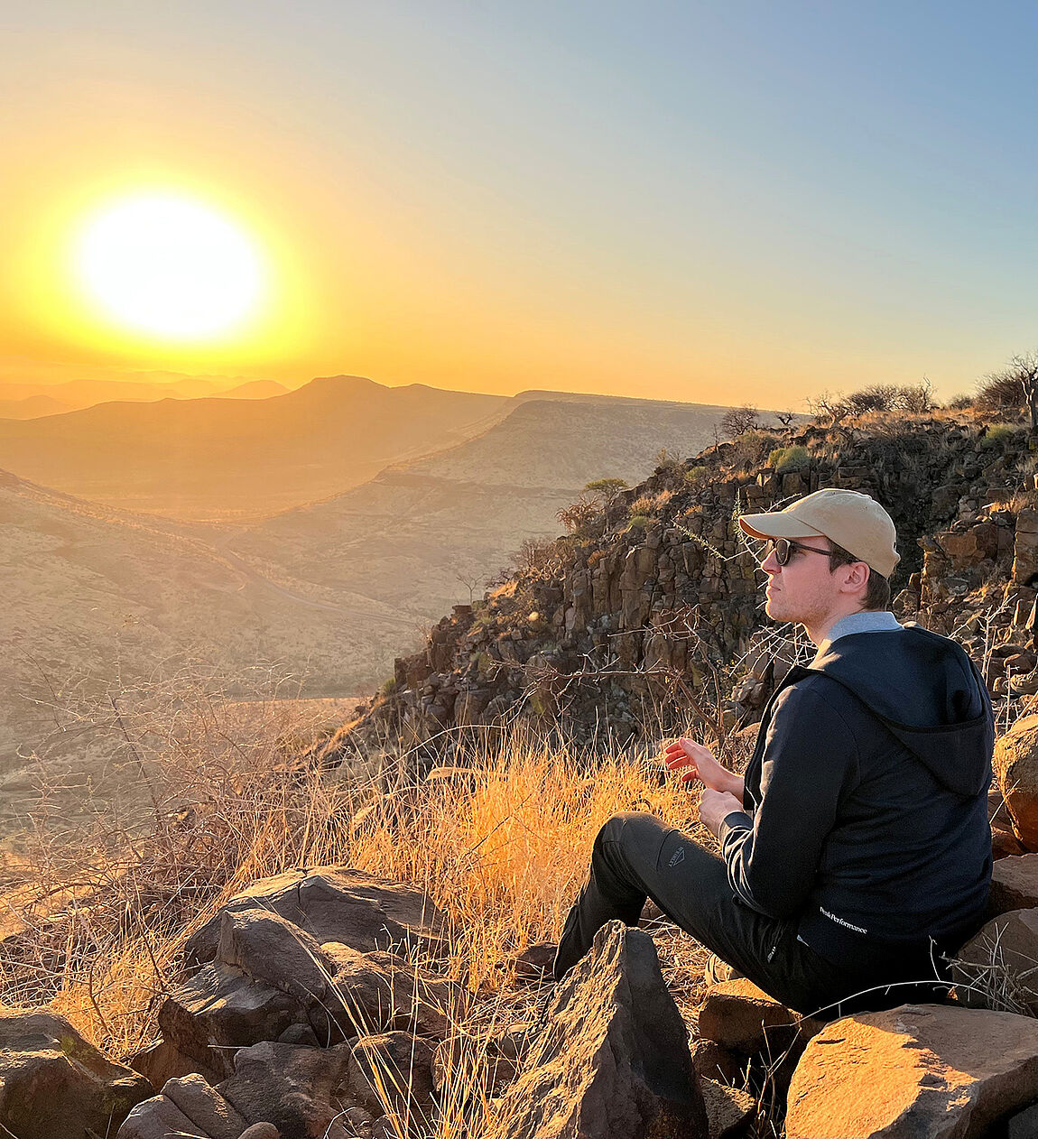 Am Rand einer Klippe sitzt ein junger Mann mit Brille und Kappe, im Hintergrund ein Tal und mehrere Hügel unter tiefstehender Sonne.