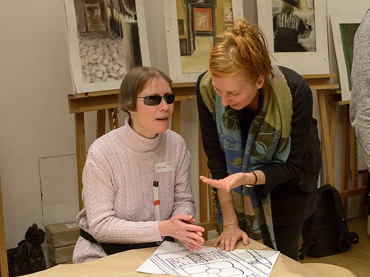 Eine Frau mit Sonnenbrille und weißem Stock sitzt an einem Tisch, der taktile Plan vor ihr wird von einer Frau beschrieben, die neben ihr steht.
