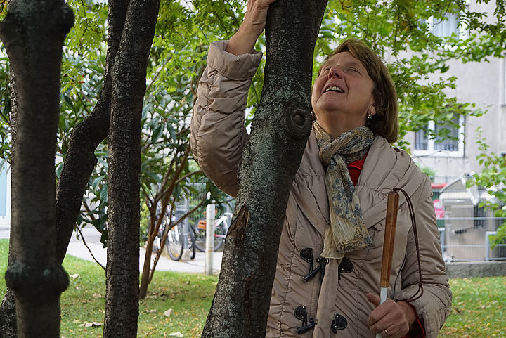 Eine Frau mit weißem Stock in der Hand ertastet mit der anderen Hand lächelnd die Rinde an einem Baumstamm in einem begrünten Hof, ihr Kopf ist nach oben gerichtet.