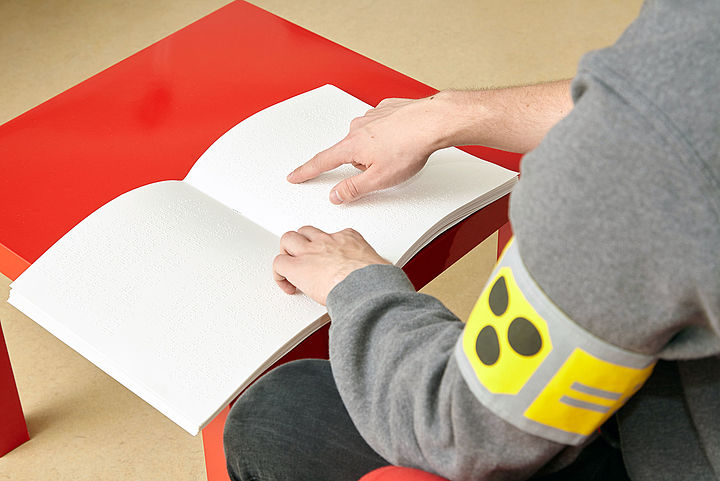 Auf einem roten Tisch liegt ein dickes weißes Buch, man sieht die Arme eines Mannes mit einer Blindenschleife links, der Zeigefinger der rechten Hand streicht über die erhabenen Braillepunkte auf der rechten Seite des Buches