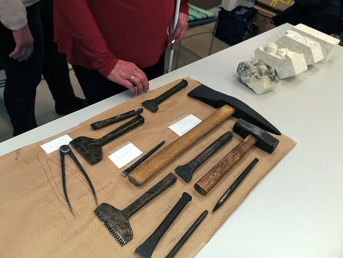 Auf einem Tisch sind knapp ein Dutzend Werkzeuge wie Hammer oder Metallkeile nebeneinander aufgelegt, eine Hand tastet danach.