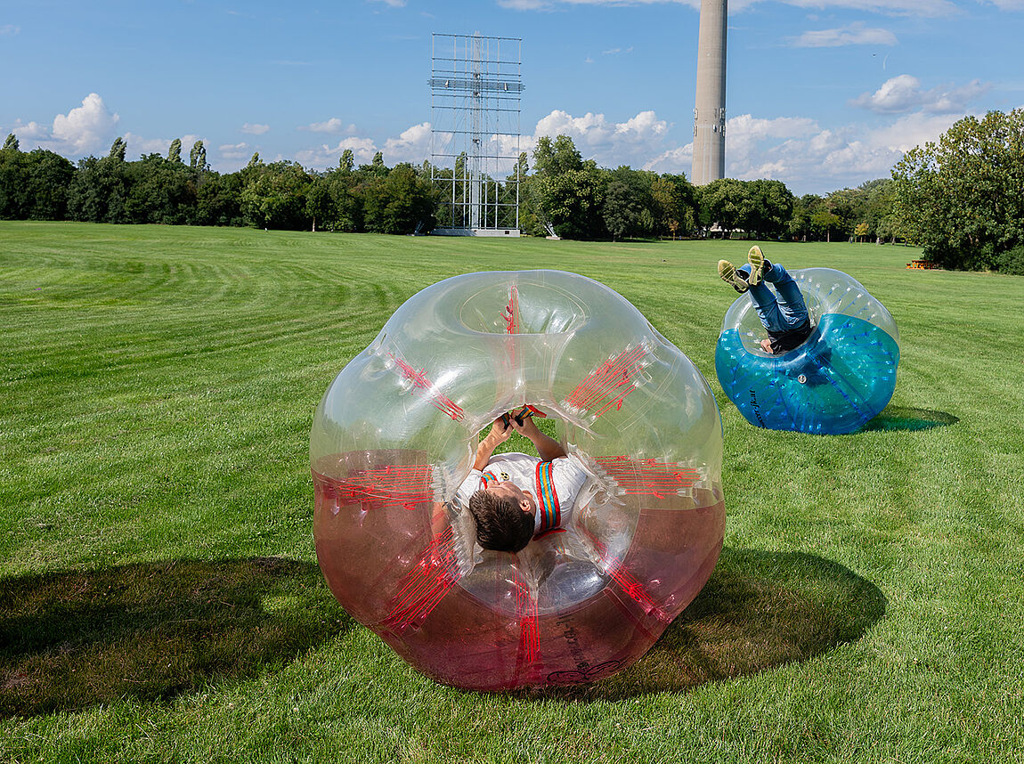 Zwei Jugendliche, jeweils in einem riesigen Ball festgeschnallt, rollen nach einer Kollision über die Wiese.