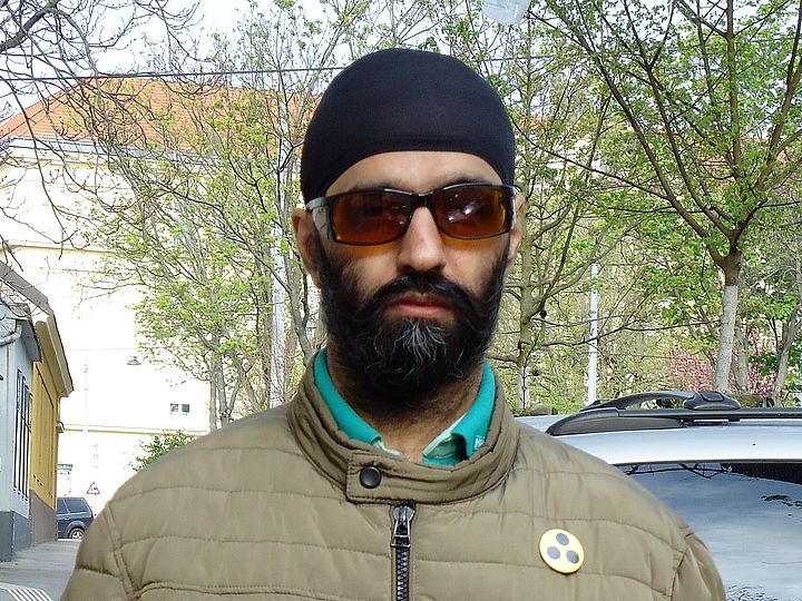 Kopf- und Schulteraufnahme eines Mannes mit Kantenfilterbrille und 3-Punkte-Button, im Hintergrund Bäume an einer Straße