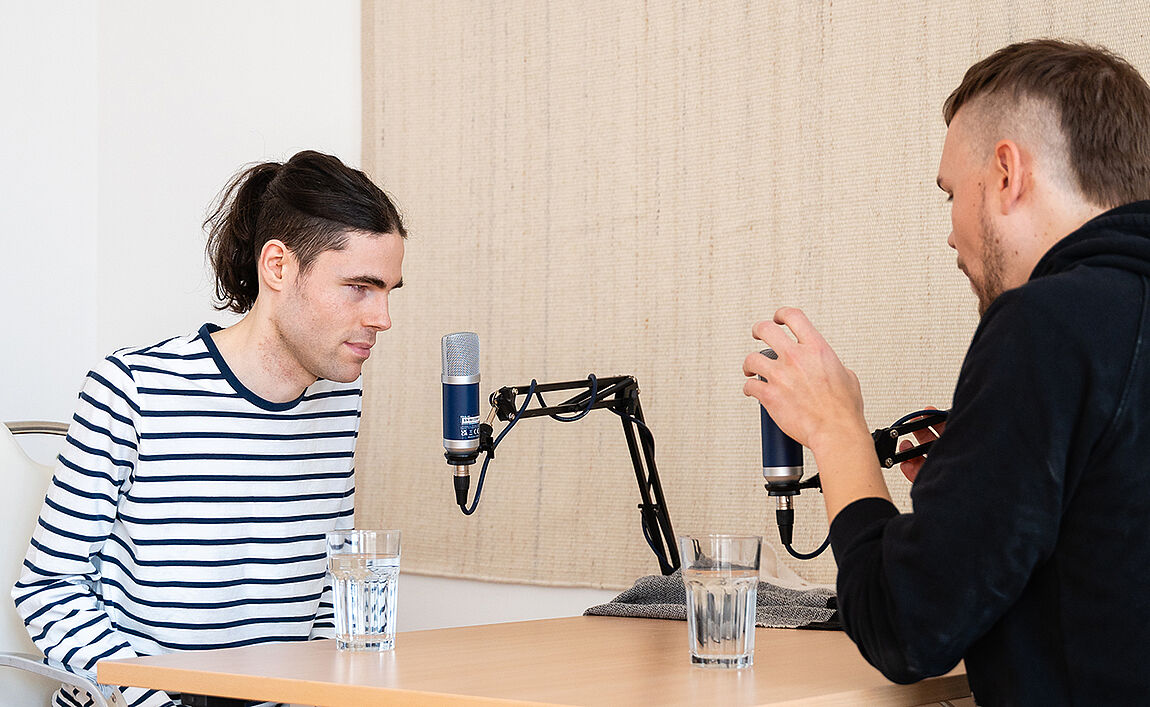 Zwei junge Männer sitzen gegenüber an einem Tisch, beide haben jeweils ein Mikrofon an einer Art Schwenkarm vor sich.