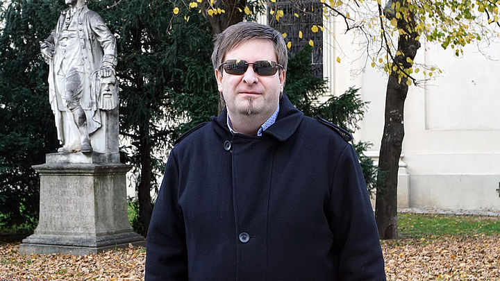 Ein Mann mit Sonnenbrille und Herbstmantel steht in einem Parkareal vor einer Statue.