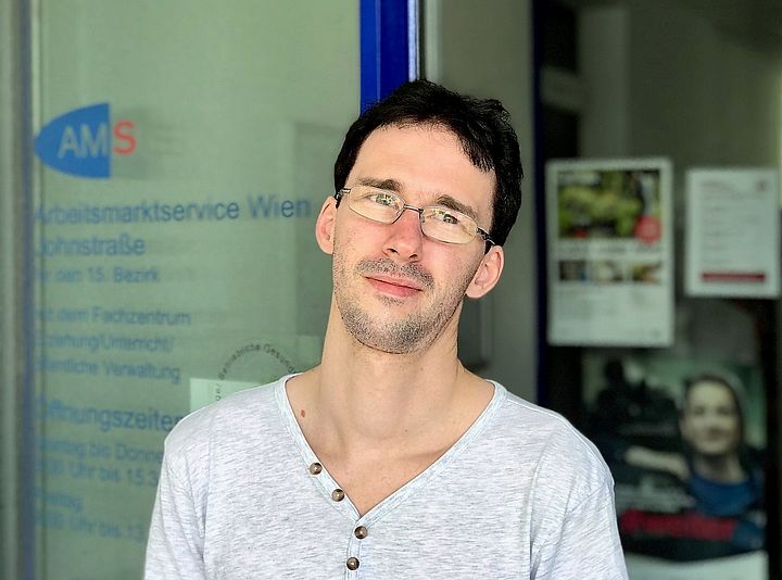 Ein junger Mann mit Brille steht vor einer Glastüre mit der Aufschrift "Arbeitsmarktservice Wien"