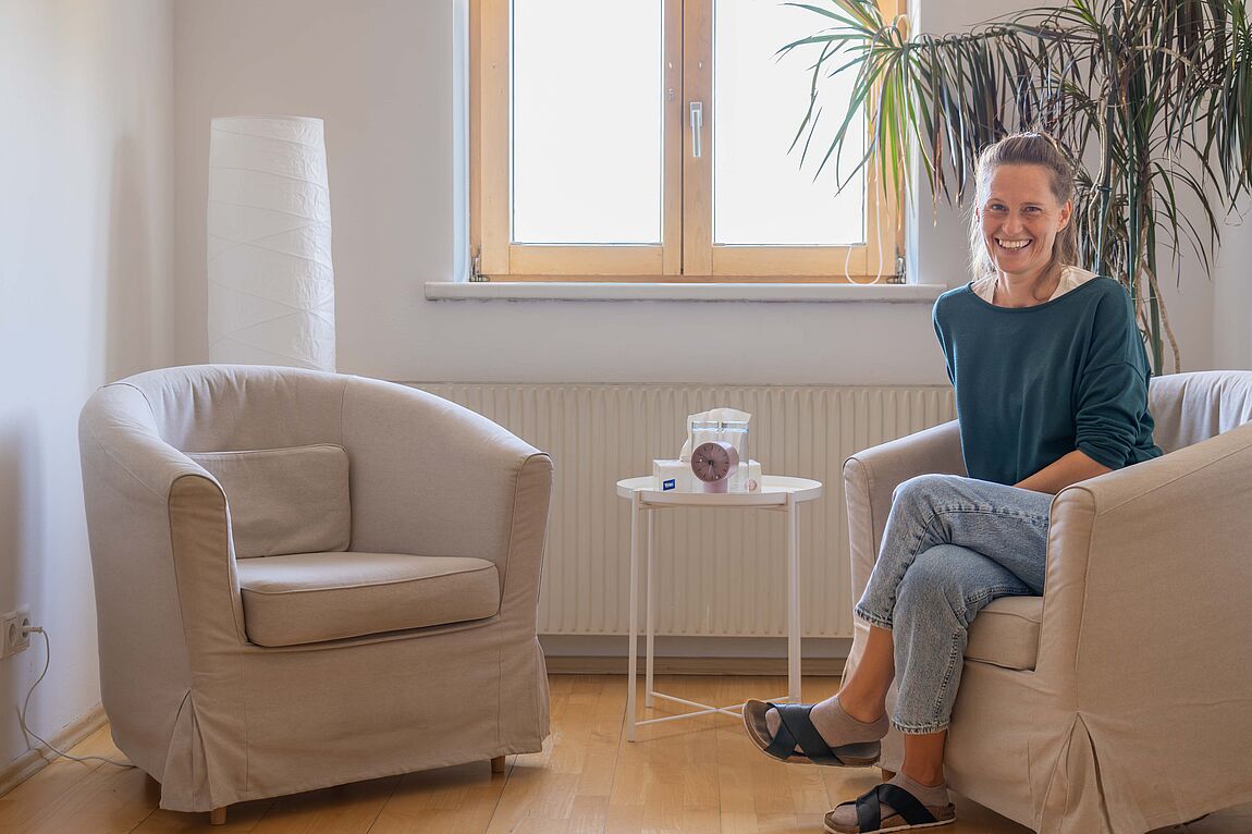 Frau sitzt lächelnd auf einem beige bezogenen Sessel in einem hellen Raum mit einem kleinen runden Tisch und einer großen Pflanze im Hintergrund