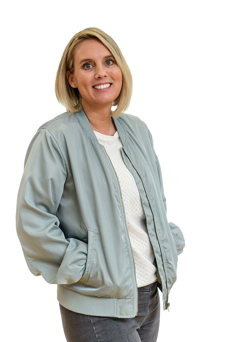Eine lächelnde Frau mit blonden Haaren in einer hellblauen Bomberjacke vor einem weißen Hintergrund.