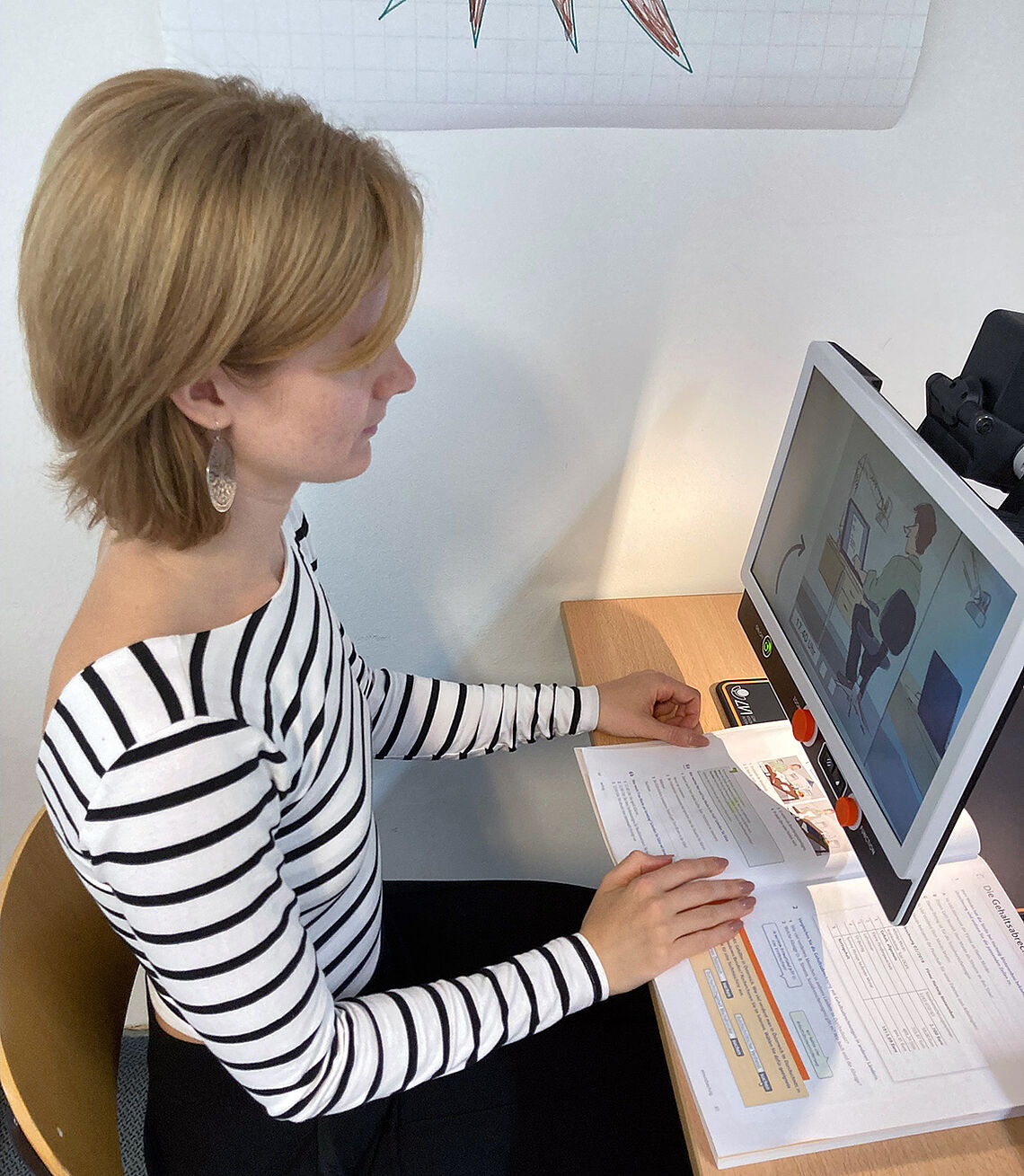 Eine junge Frau mit halblangen Haaren sitzt beim Arbeitstisch vor einem Monitor, auf dem die Buchseiten angezeigt werden, die unter dem Monitor offen liegen.