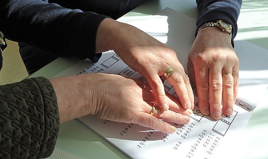 Nahaufnahme von drei Händen, die Finger ertasten die Position von Braille Schriftzeichen in einer Broschüre
