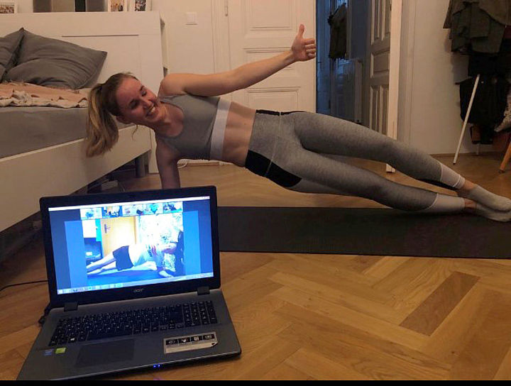 Auf dem Boden einer Wohnung ein Laptop, auf dessen Bildschirm jemand eine Turnübung vorzeigt, dahinter Melanie auf einer Bodenmatte in seitlich abgestützter, gestreckter Körperhaltung, mit der freien Hand streckt sie den Daumen lachend nach oben.