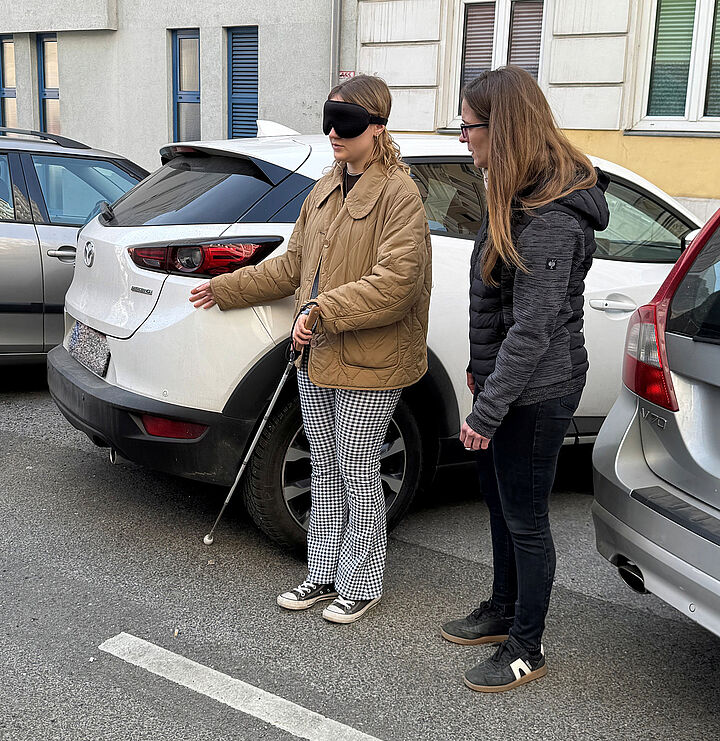 Eine Frau mit Augenbinde und weißem Stock beim Heck eines geparkten Autos, eine weitere Frau steht daneben und beobachtet.