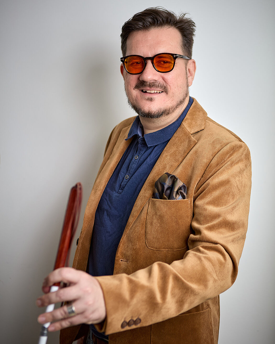 Ein Mann mit orange getönter Sonnenbrille, Hemd und Sakko, in der nach vorne ausgestreckten Hand ein Weißer Stock.