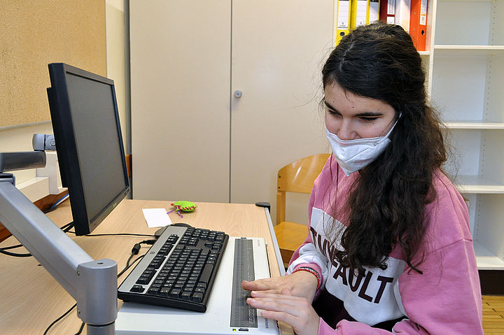 Eine Schülerin mit langen, dunklen Haaren, einer FFP2-Maske und einem pinken Sweater sitzt vor einer Computertastatur mit vorgelagerter Braillezeile.
