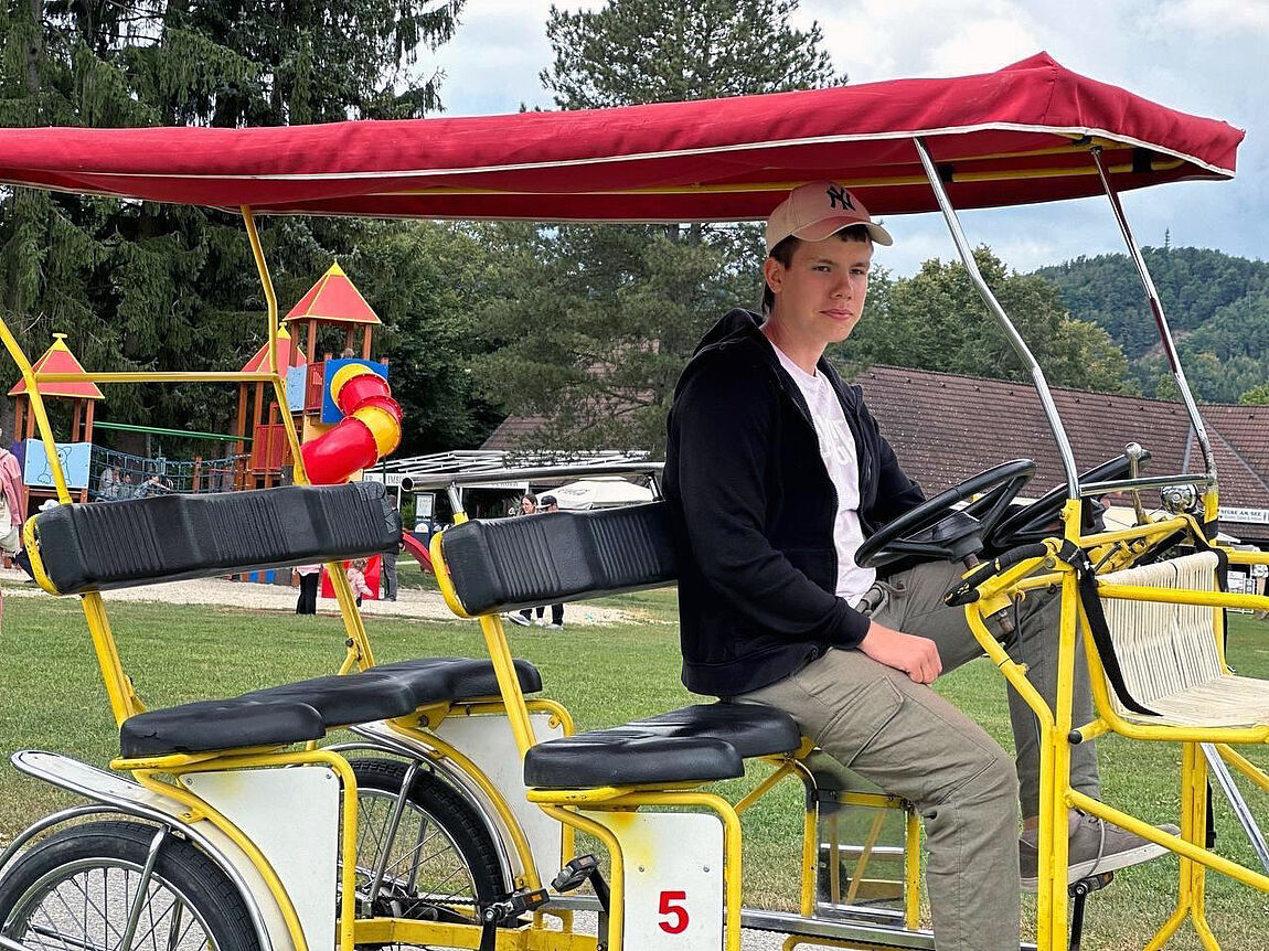 Ein junger Mann auf der vorderen von zwei Bankreihen am Steuer einer überdachten Rikscha vor einem Spielplatz.