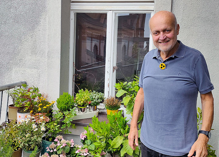 Bernhard Ludwig steht neben Blumenstauden auf dem Balkon vor einem Fenster, er trägt ein Kurzarm-Shirt und einen Button mit drei schwarzen Punkten auf gelbem Hintergrund.