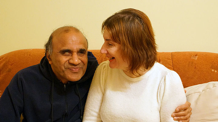 Mahendra in einem schwarzen Sweater legt seinen Arm um Adriana, die einen weißen Pullover trägt; sie sitzen nebeneinander auf einem orangen Sofa und lachen