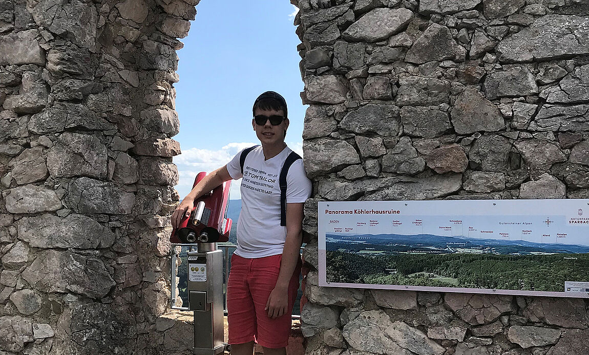 Ein junger Mann mit Sonnenbrille vor einem Fenstererker in einer Burgmauer, neben ihm ein Schild mit Details zum "Panorama Köhlerhausruine".