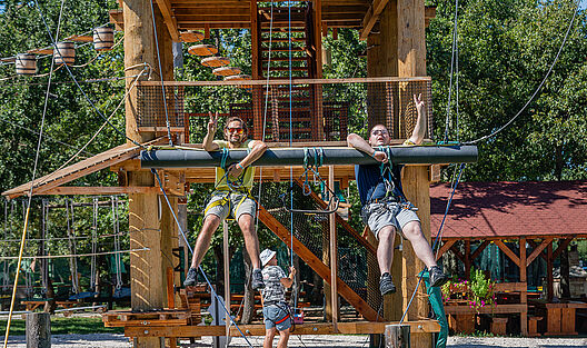 Bei strahlender Sonne hängen ein junger Teilnehmer und ein Betreuer festgegurtet an einer Riesenschaukel in luftiger Höhe im Kletterpark und zeigen dabei fröhlich das Peace-Zeichen in die Kamera.