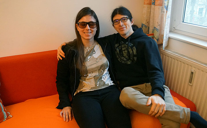 Eine junge Frau mit Sonnenbrille und ein junger Mann mit Brille lächelnd und einander umarmend auf einem Sofa sitzend, beide haben lange dunkle Haare.