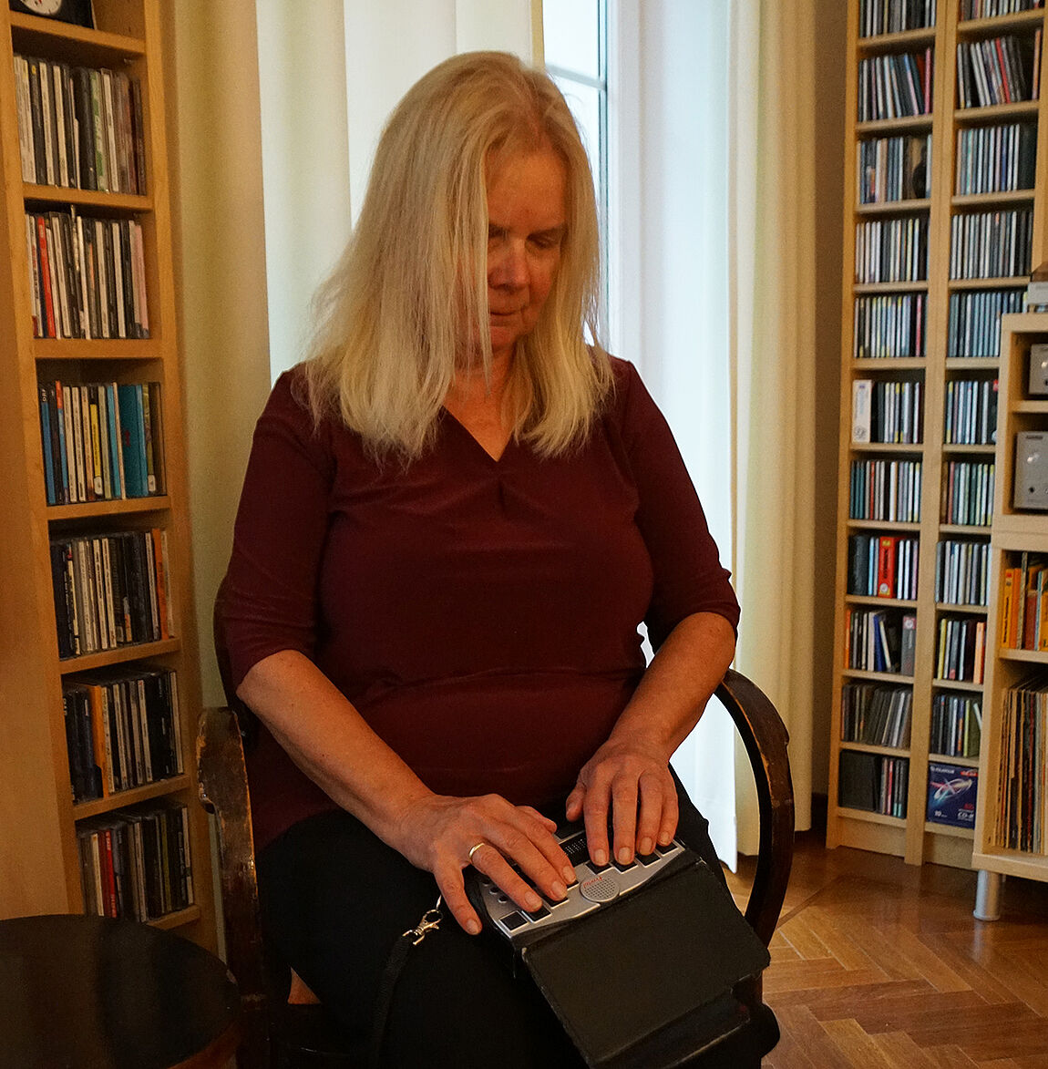 Eine Frau mit langen hellblonden Haaren sitzt vor Regalwänden auf einem Sessel. Auf ihrem Schoß ein technisches Gerät in der Größe eines Buches, sie tippt mit Fingern beider Hände auf große Tasten.
