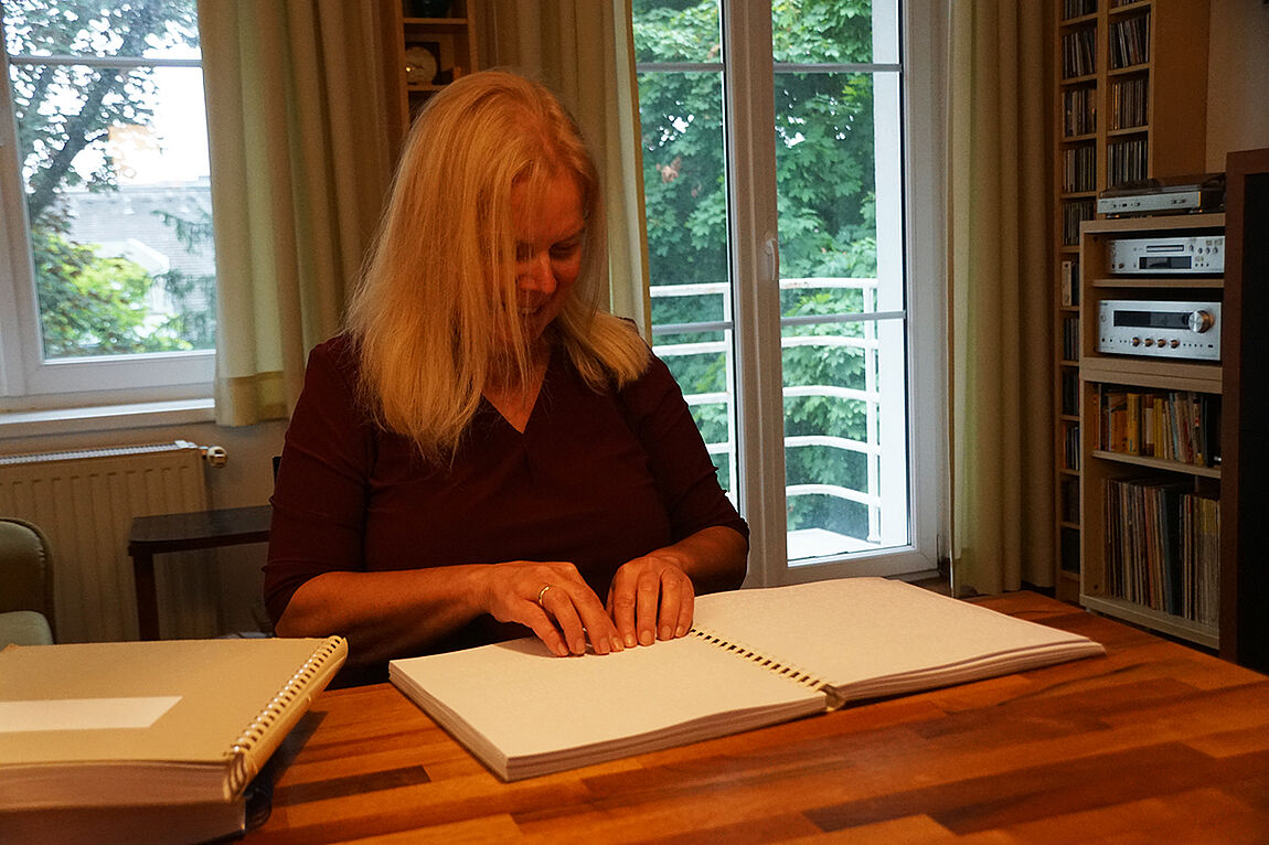 Eine Frau mit langen hellblonden Haaren sitzt an einem Tisch, vor ihr ein dickes Heft in Brailleschrift. Sie liest mit Fingern beider Hände darin.