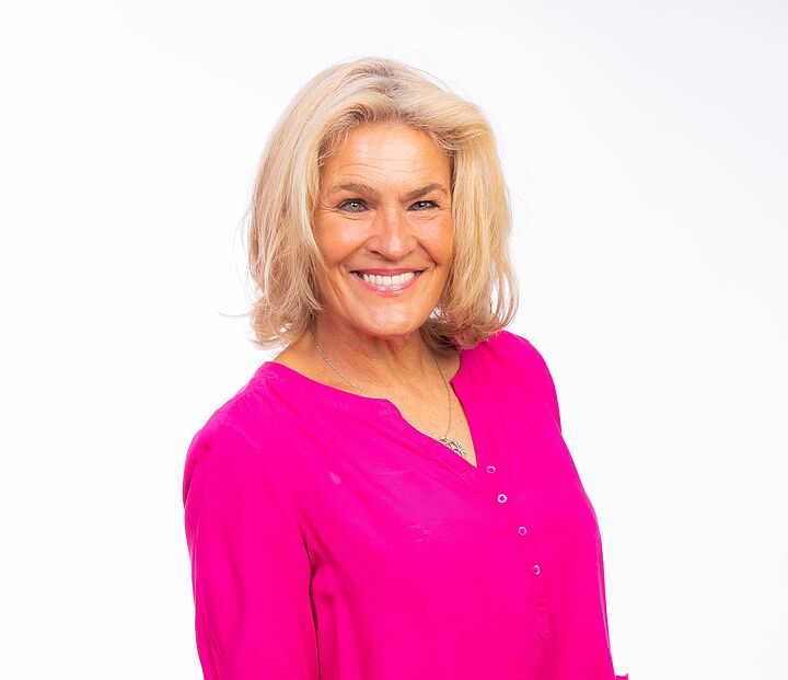 Vor weißem Hintergrund ein Portraitfoto einer lachenden blonden Frau mit pinker Bluse.