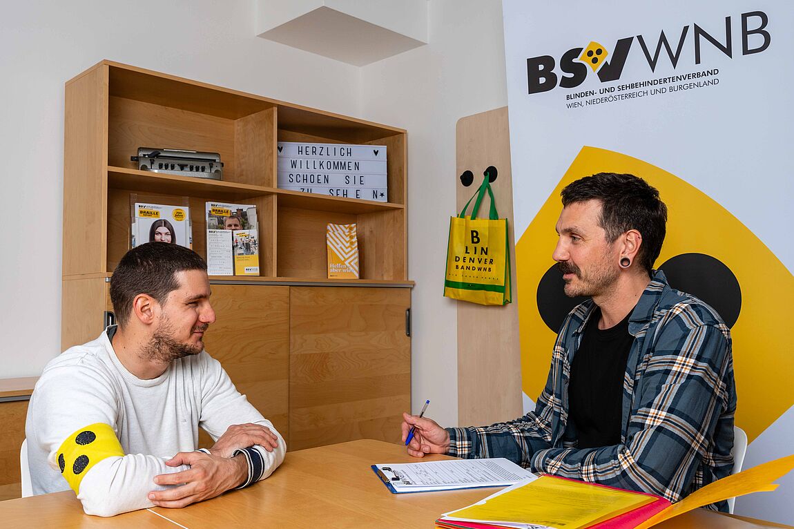 2 Personen sitzen sich an einem Tisch gegenüber. Eine der Personen trägt eine gelbe Schleife mit 3 schwarzen Punkten. Beide haben ein Lächeln im Gesicht.. Im Hintergrund steht ein rollup mit dem Logo des Blindenverbandes.