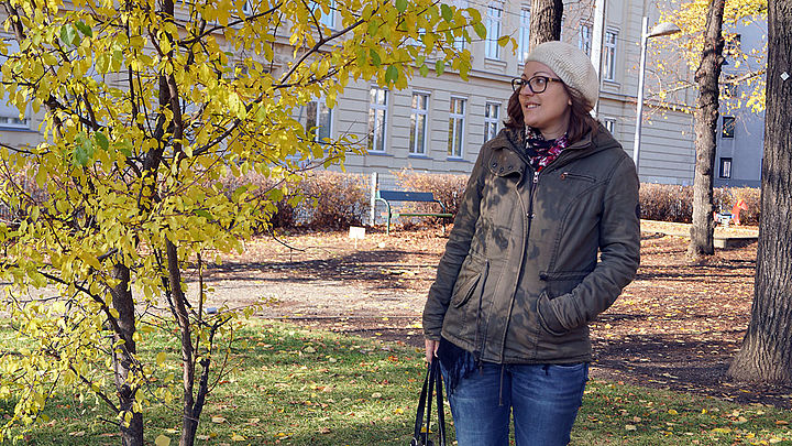Eine Frau mit warmer Jacke und Haube steht an einem sonnigen Herbsttag in einem Park neben kleinen Bäumen mit gelben Blättern.