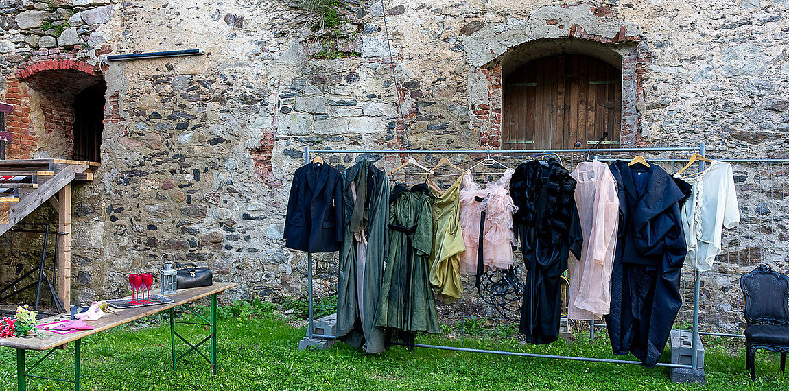 Vor den Burgmauern ein langer Kleiderständer mit diversen Kostümen, links davor ein Tisch mit Requisiten, beispielsweise Gläser oder eine Laterne.