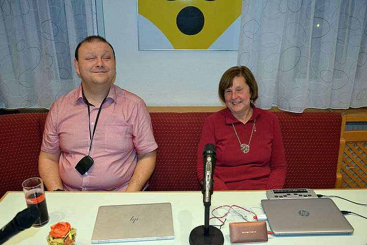 Ausgerüstet mit Notebooks und technischen Hilfsmitteln sitzen Bayram und Eva lachend beim Vortragstisch in den Louis Braille Stuben, im Hintergrund Vorhänge und das BSVWNB Logo