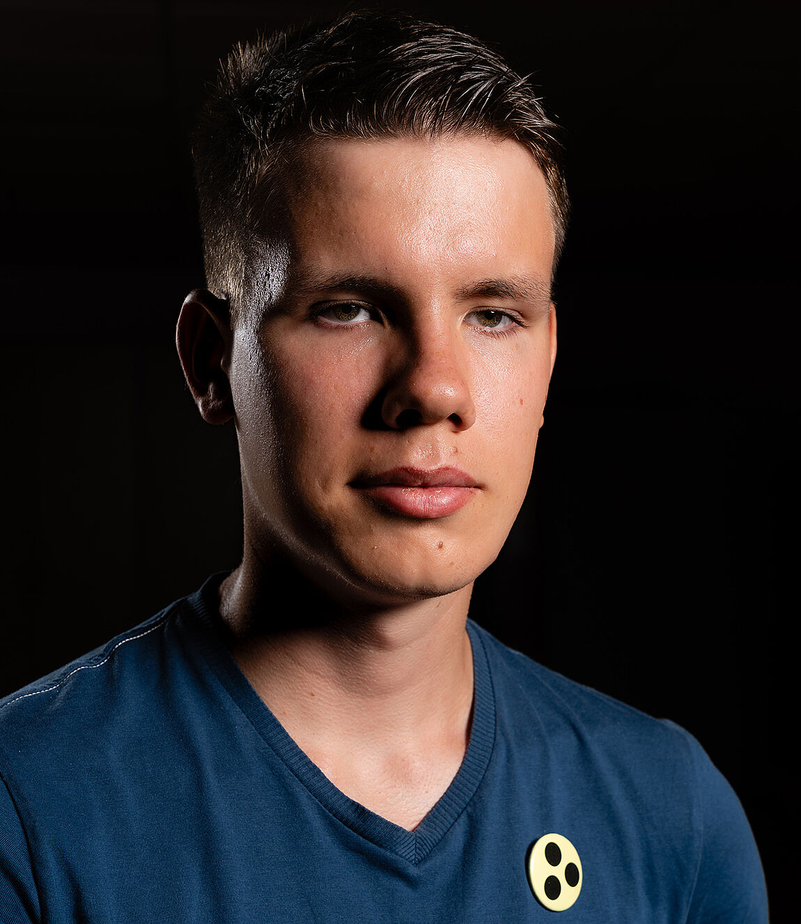 Portraitfoto eines jungen Mannes mit gelbem Ansteckbutton mit drei schwarzen Punkten, vor dunklem Hintergrund.
