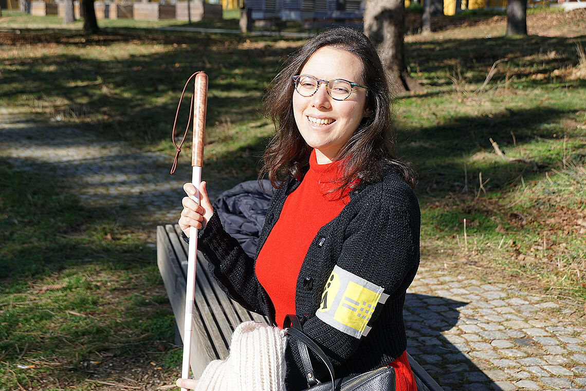 Eine lachende Frau mit schulterlangen dunklen Haaren und Brille auf einer Parkbank. Über einem roten Kleid trägt sie eine dunkle Strickweste, am linken Arm eine gelbe Schleife. In der rechten Hand ein Weißer Stock.