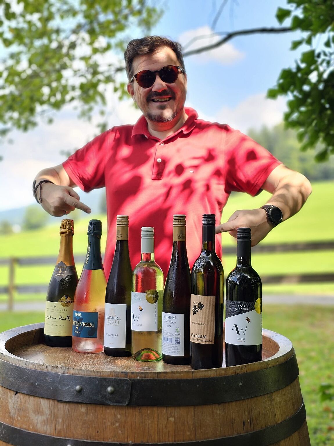 Im Schatten eines Baumes vor einer grünen Wiese zeigt ein Mann mit beiden Zeigefingern stolz auf ein Fass, auf dem mehrere Weinflaschen nebeneinander aufgestellt sind.