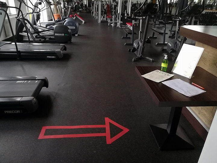 Eine große Halle mit Fitness-Geräten. Vorne links ein Laufband, am Boden zentral ein Pfeil aus rotem Klebeband, der nach rechts zum Anmeldetisch zeigt.