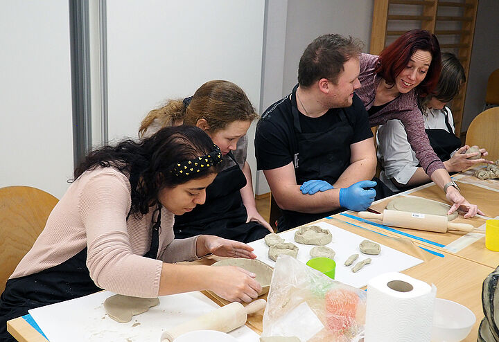 Vier Jugendliche an einem langen Tisch; auf dem Tisch vor ihnen halbfertige Ton-Werkstücke. Die Workshopleiterin beugt sich zwischen zwei Jugendlichen über den Tisch und ertastet einen Tonteller.
