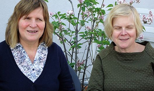 Die beiden Damen sitzen auf einem Balkon oder einer Terrasse vor einem kleinen blühenden Strauch und lächeln in Richtung Kamera