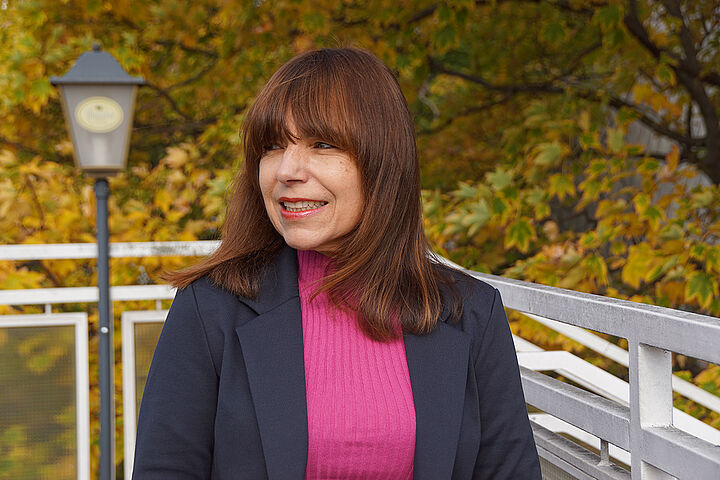 Eine Frau mit glatten langen Haaren blickt lachend zur Seite, im Hintergrund ein Geländer und Äste eines Baumes.