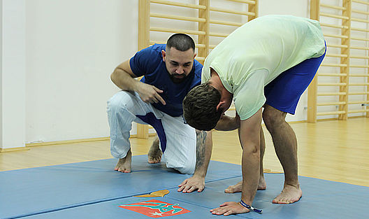 Ein Jugendlicher geht auf der blauen Matte in Position, der Lehrer kniet daneben und überwacht die Bewegung.