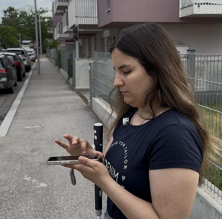 Eine junge Frau mit langen, dunklen Haaren bedient fokussiert ihr Smartphone auf einem Gehweg. In der Hand hält sie einen modernen, schwarz-weißen Langstock. Ihre Haltung strahlt Kompetenz und Unabhängigkeit im urbanen Raum aus, im Hintergrund sind moderne Wohngebäude und parkende Autos zu sehen.