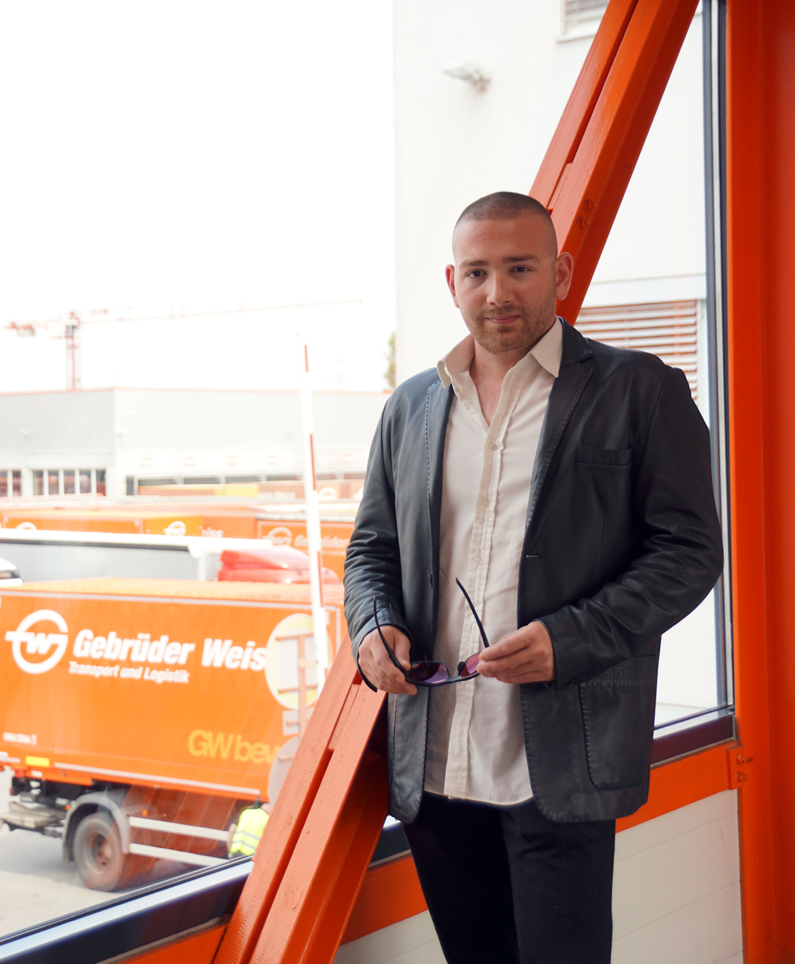 Ein junger Mann vor einem großen Fenster, im Außenbereich dahinter mehrere Lastwägen.