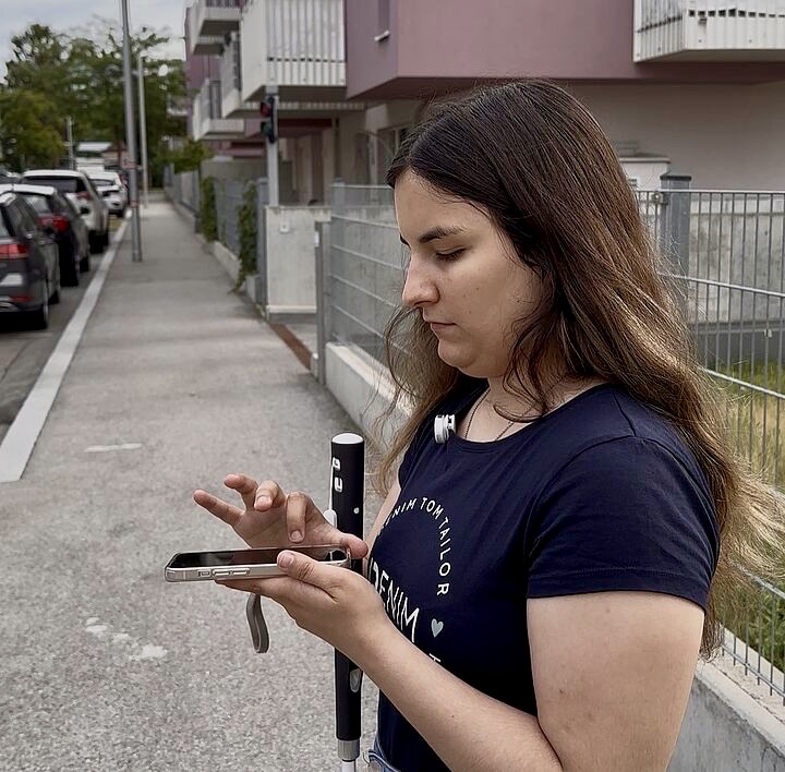 Eine junge Frau mit langen, dunklen Haaren bedient fokussiert ihr Smartphone auf einem Gehweg. In der Hand hält sie einen modernen, schwarz-weißen Langstock. Ihre Haltung strahlt Kompetenz und Unabhängigkeit im urbanen Raum aus, im Hintergrund sind moderne Wohngebäude und parkende Autos zu sehen.
