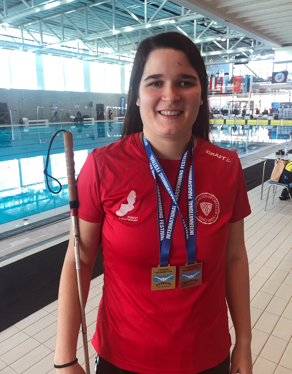 In Sportmontur, mit weißem Langstock, und zwei Medaillen um den Hals steht eine junge Frau lächelnd in einer großen Schwimmhalle.