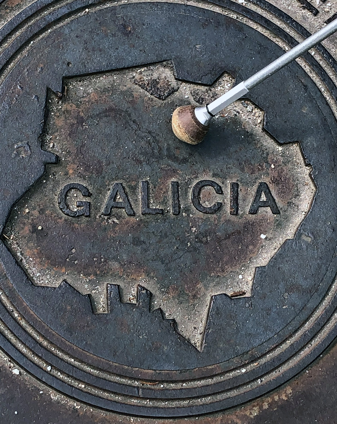 Die Spitze eines weißen Langstocks bei einem Kanaldeckel mit der Aufschrift: Galicia