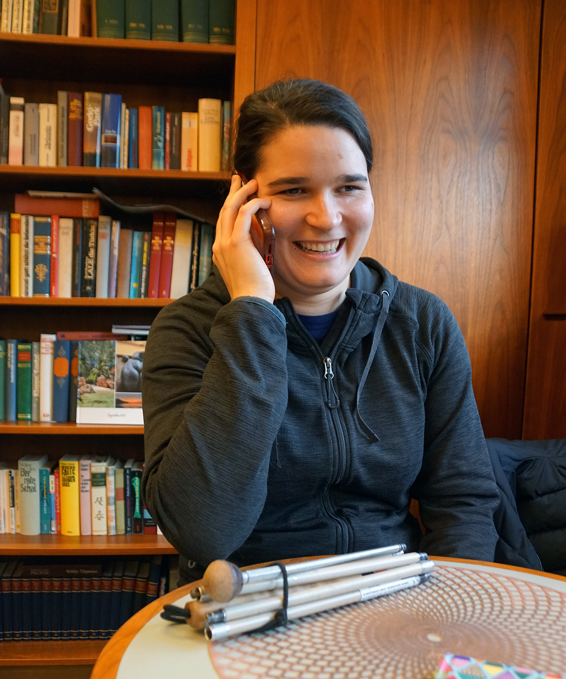 Eine junge Frau telefoniert am Handy, ein zusammengeklappter Langstock liegt vor ihr auf dem Tisch. Im Hintergrund ein dicht befüllter Bücherschrank.