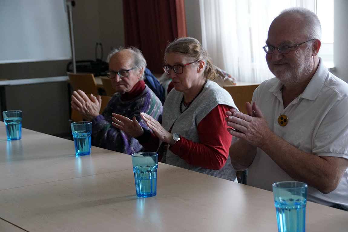 Drei Personen nebeneinander sitzend an einem Tisch, sie klatschen und klopfen mit den Händen.